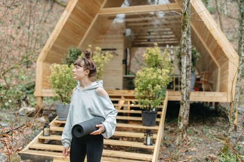 Free Woman holding a yoga mat outside a wooden cabin in a forest, ready for a wellness retreat. Stock Photo