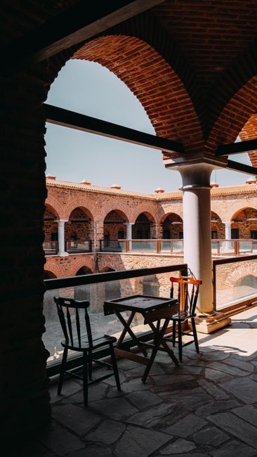 Table with chairs placed near glass fence under roof of aged stone house with arched passages on sunny day in town
