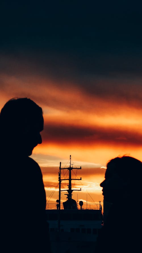 Silhouettes side view of anonymous couple standing near ship against colorful cloudy sky at sunset time in port in nature