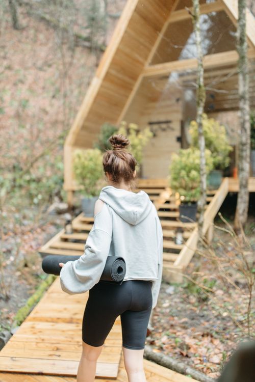 Free A woman carrying a yoga mat approaches a modern wooden cabin in a serene forest setting, depicting a tranquil lifestyle. Stock Photo