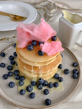 A stack of pancakes topped with blueberries and pink cotton candy, paired with syrup.