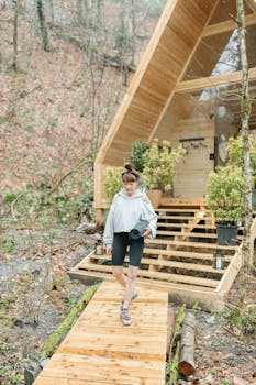 Woman in casual attire walking on a wooden boardwalk outside a modern cabin surrounded by nature.