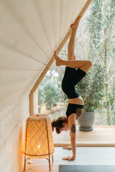 A woman performs a yoga handstand against a wall inside a cozy wooden room.