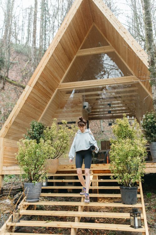 Free Woman in casual sportswear descends wooden cabin stairs surrounded by green plants. Stock Photo