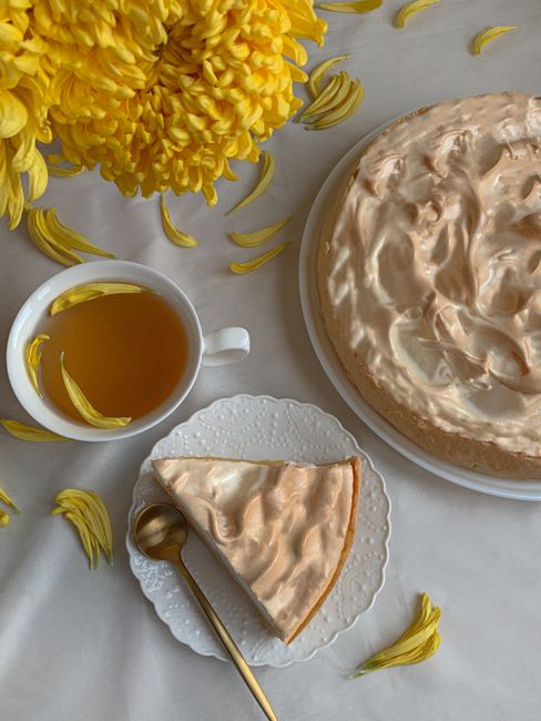 A slice of lemon meringue pie with a cup of tea and yellow chrysanthemums.