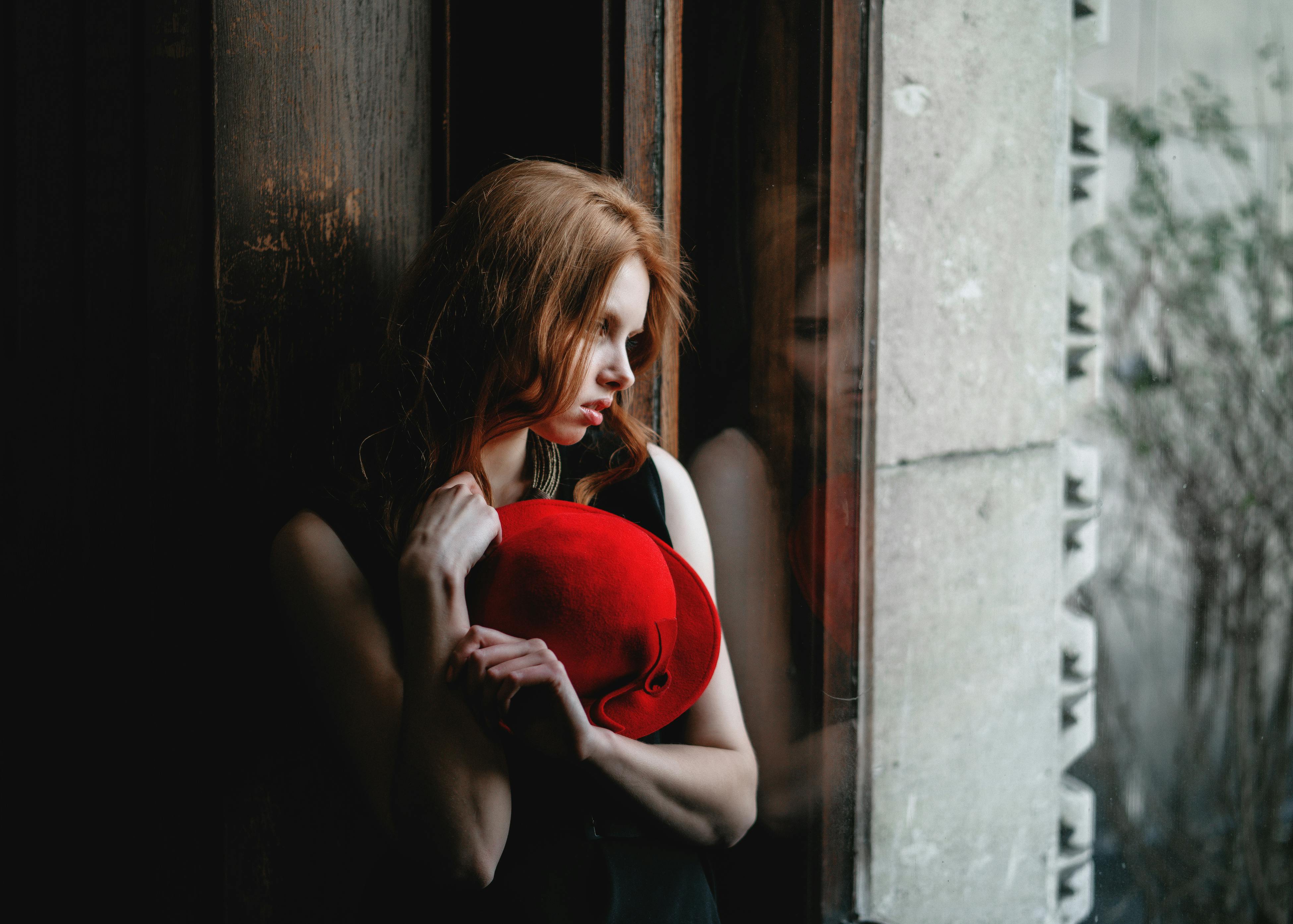 A contemplative woman stands by a window, holding a vibrant red hat, reflecting introspective mood.
