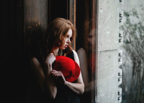 Free A contemplative woman stands by a window, holding a vibrant red hat, reflecting introspective mood. Stock Photo