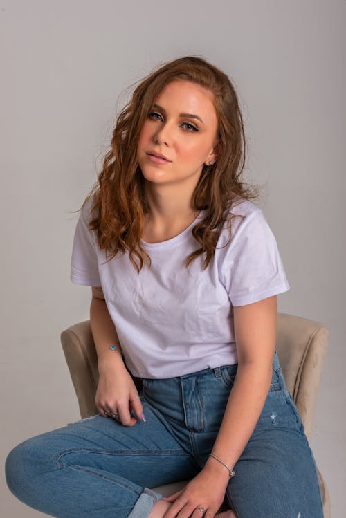 Woman in White Crew Neck T-shirt and Blue Denim Jeans Sitting on a Chair