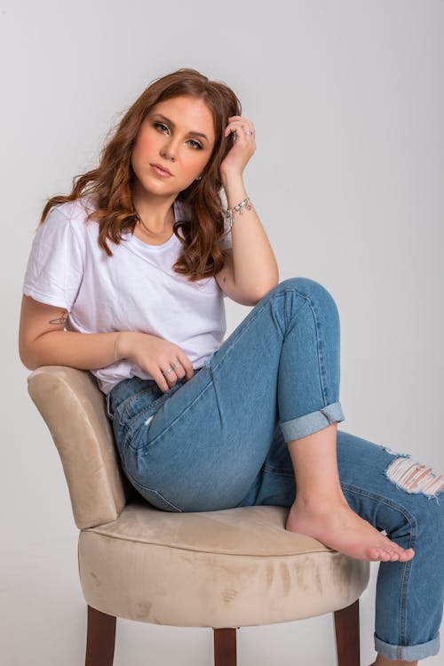 Woman in White Crew Neck T-shirt and Blue Denim Jeans Sitting on a Chair