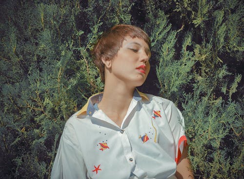 Mindful young female with short hair in stylish blouse with fish ornament against green shrub in park