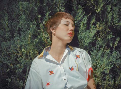 Mindful young female with short hair in stylish blouse with fish ornament against green shrub in park