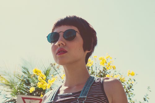 Free Trendy young female with short brown hair in sunglasses looking forward against blossoming flowers in sun ray Stock Photo