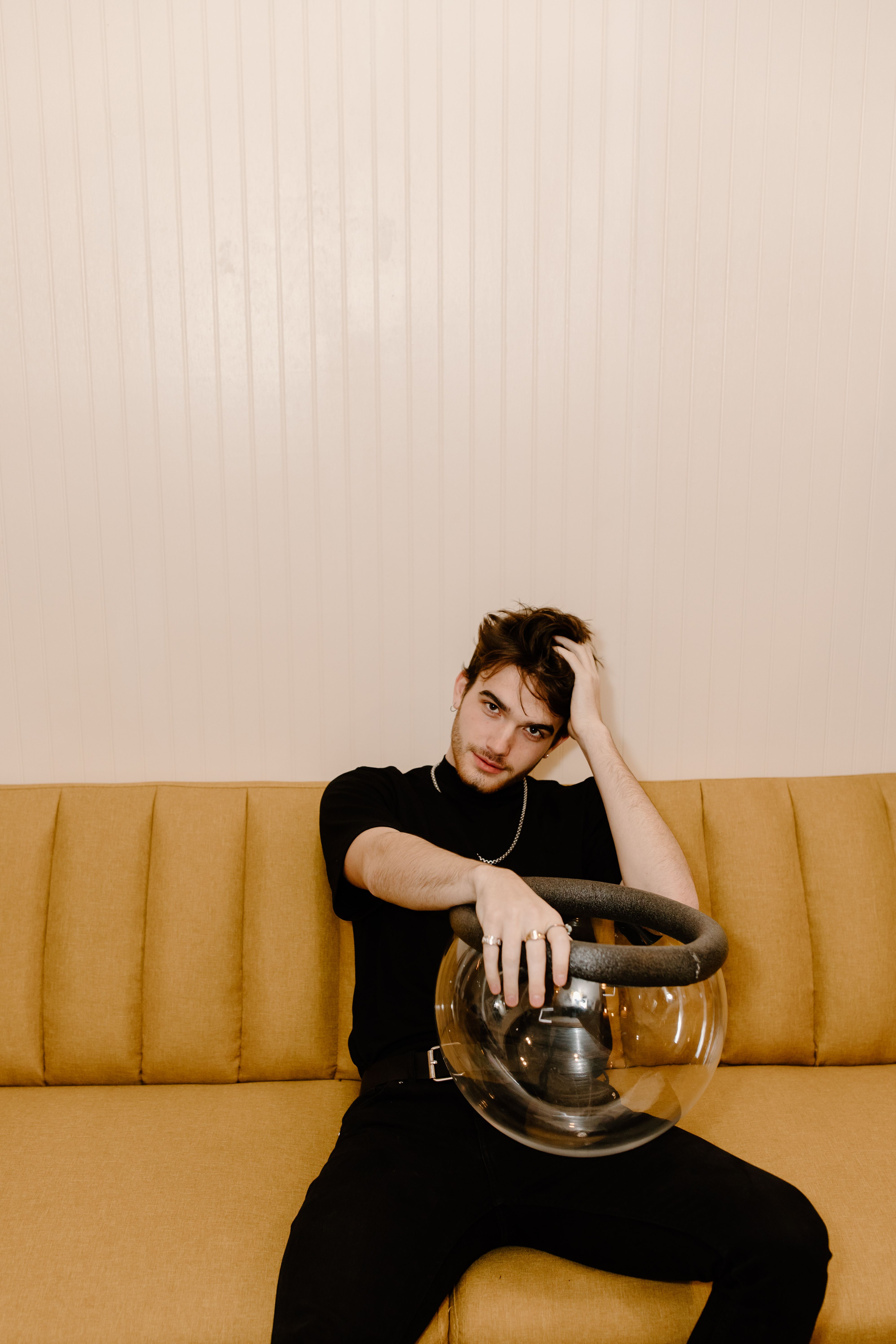 Stylish man seated indoors with a futuristic helmet, modern and introspective pose.