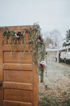A rustic wooden door decorated with flowers and greenery serves as an outdoor wedding entrance.