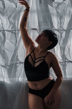 Elegant woman in black lingerie posing against a sheer curtain, showcasing confidence and style.