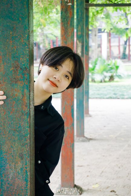 Smiling young woman peeking from behind a rusty column outdoors in a park setting.