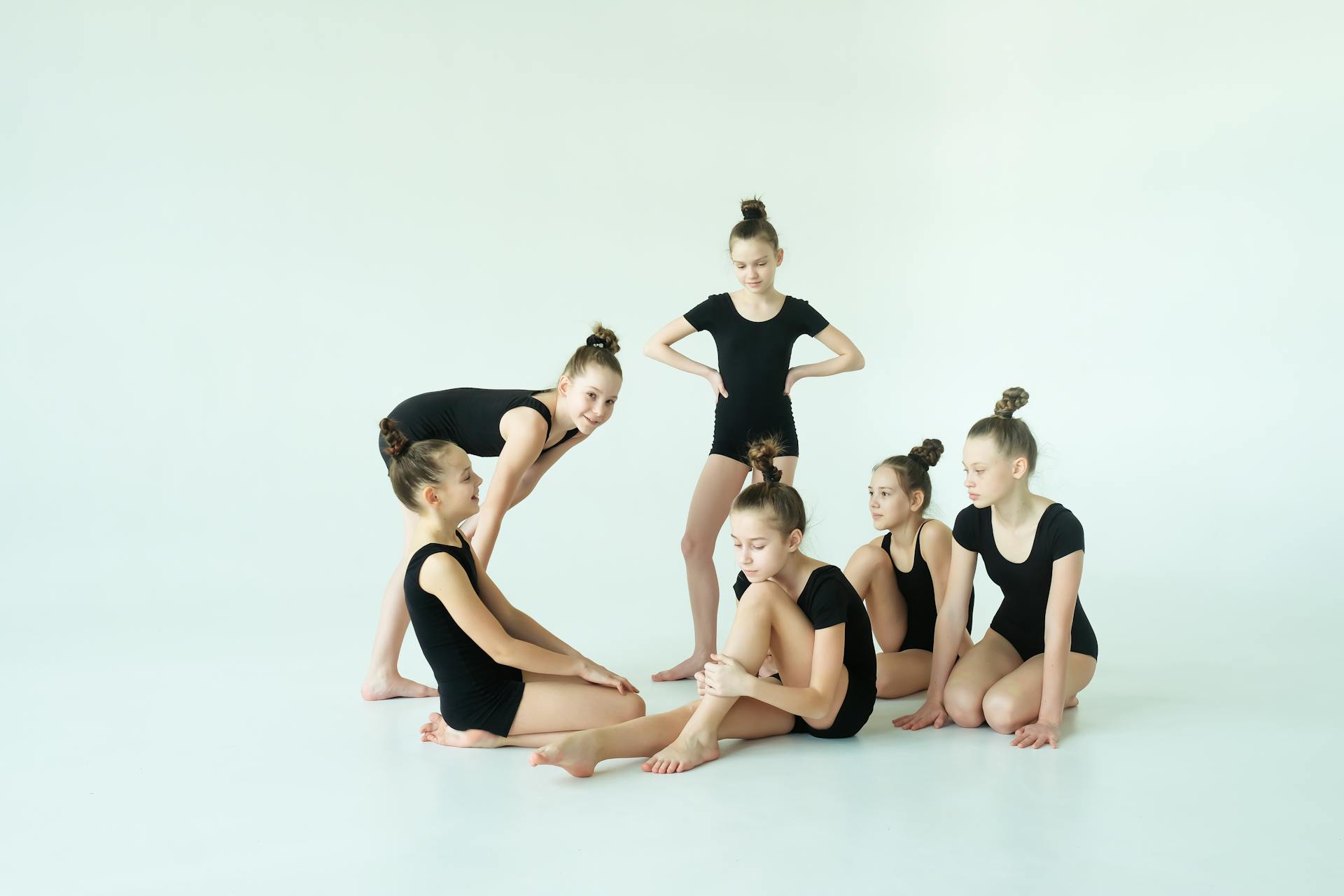 Six young girls in black leotards practicing in a bright studio.