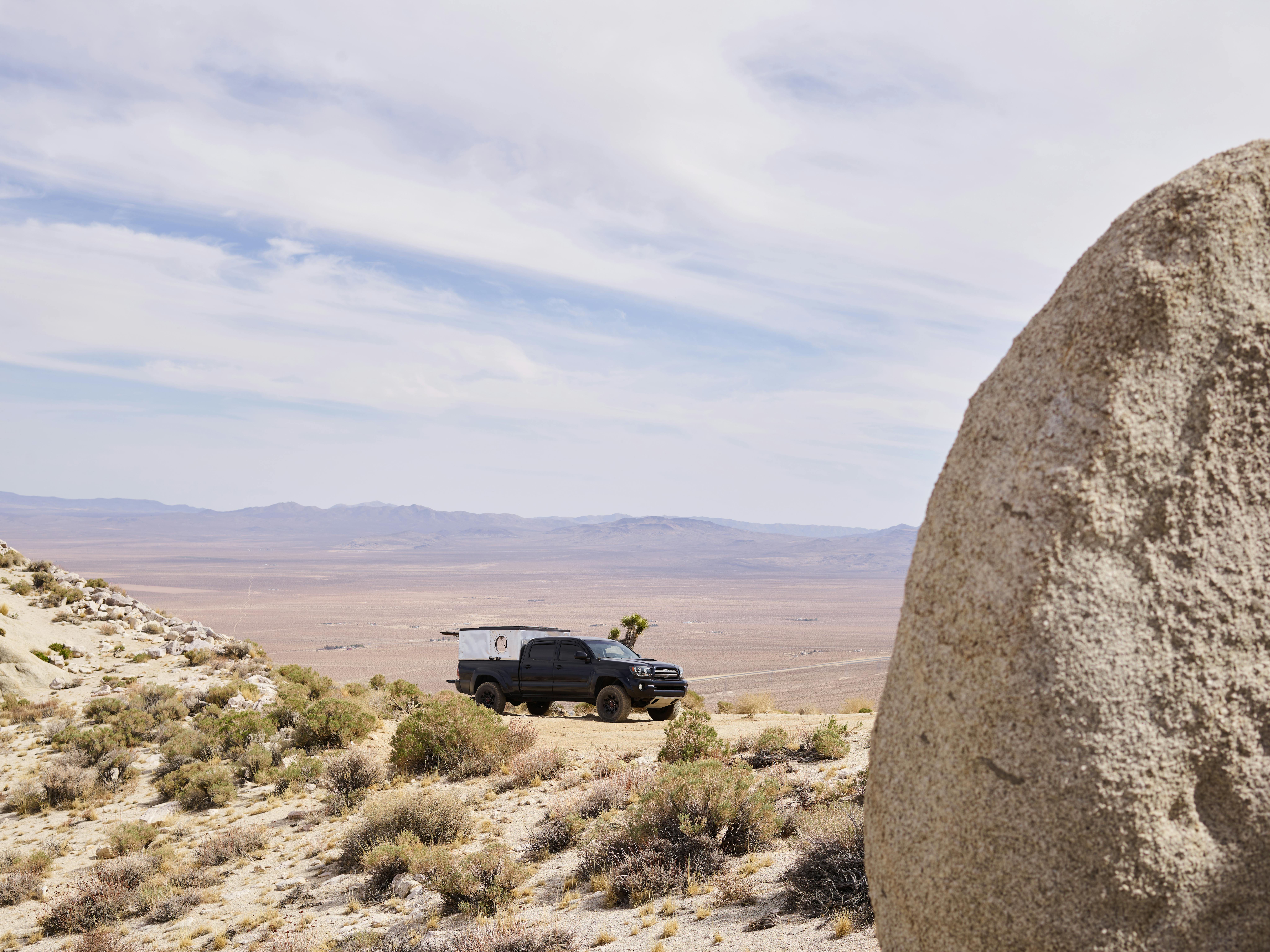 A rugged pick-up truck traverses a vast desert landscape under a clear sky, surrounded by arid terrain.