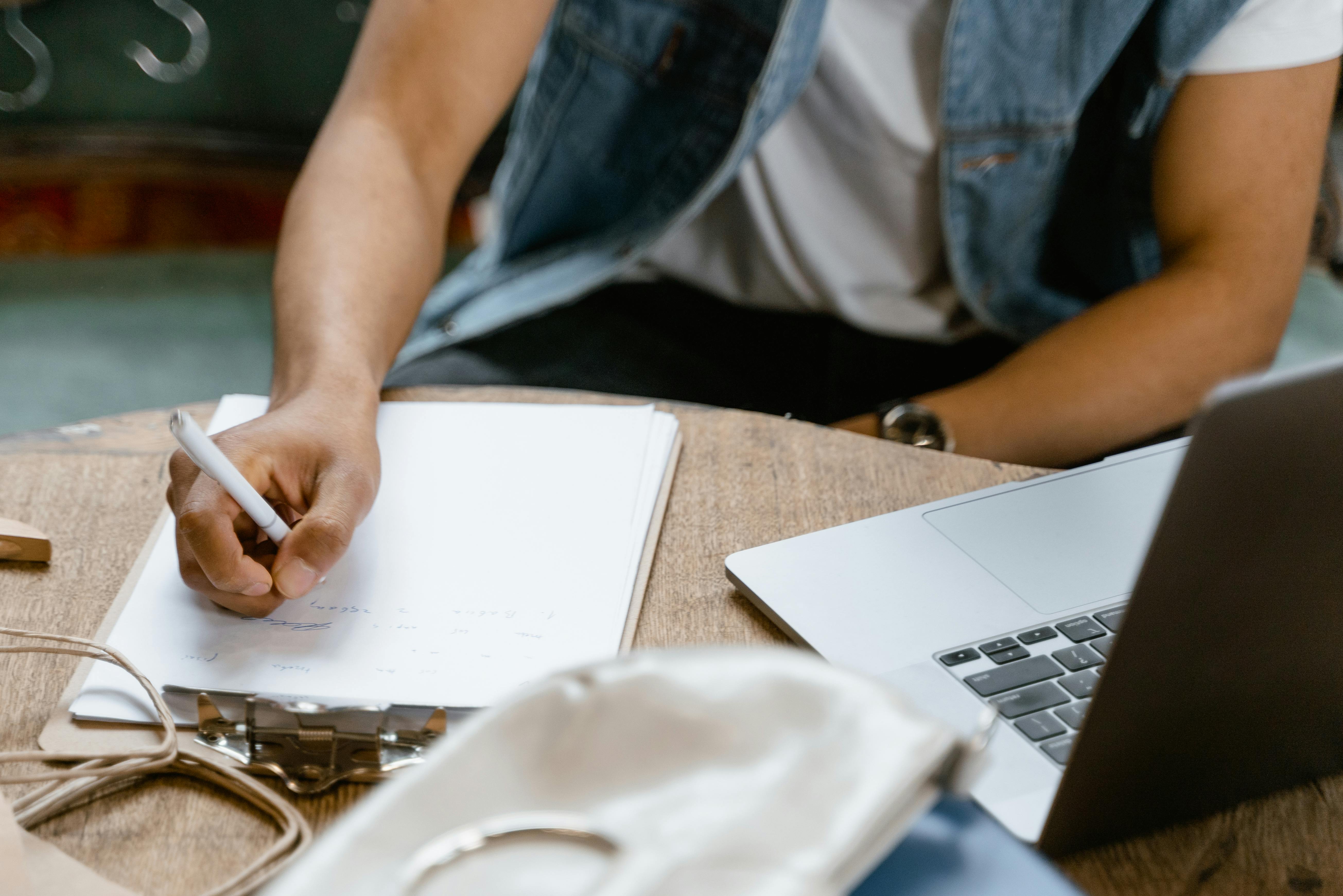 A person in denim vest writing notes on clipboard by laptop, indoors.