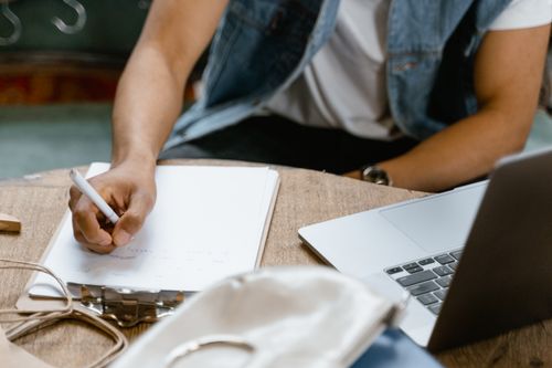 Free A person in denim vest writing notes on clipboard by laptop, indoors. Stock Photo