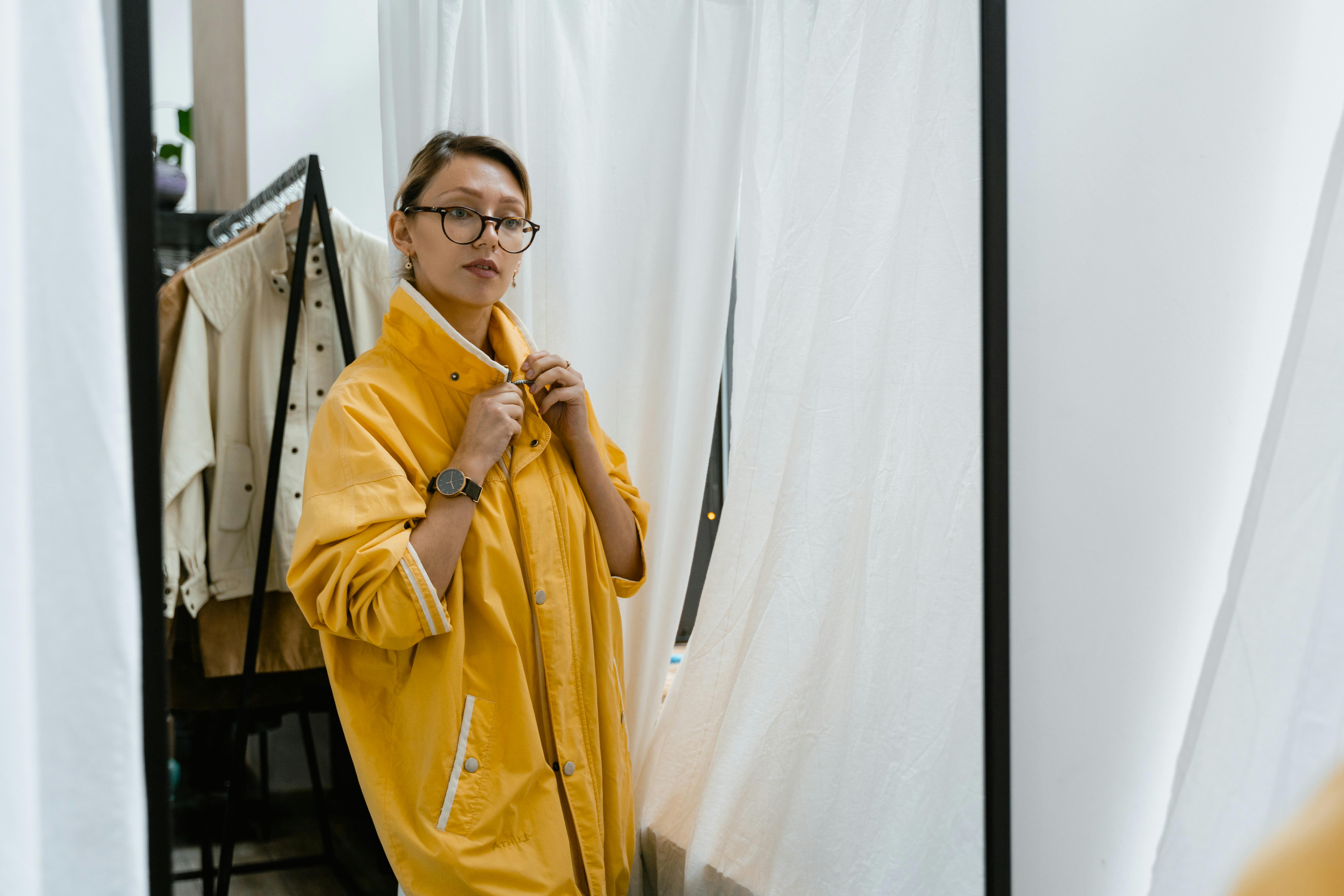 A woman in a yellow coat checks her look in a boutique fitting room mirror.