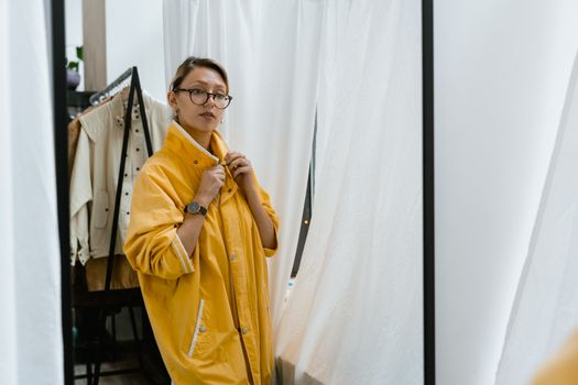 A woman in a yellow coat checks her look in a boutique fitting room mirror.