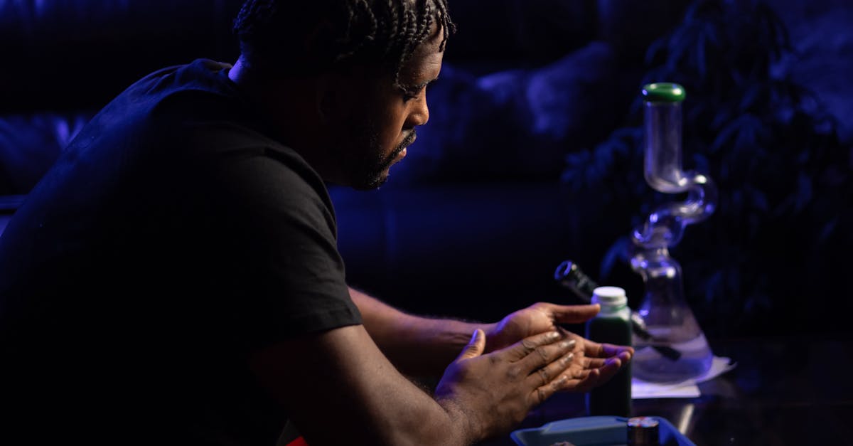 A man rolls cannabis in a dimly lit room, surrounded by smoking paraphernalia.