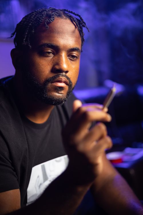 Free A thoughtful man holding a smoke in a dim, relaxed setting with a background hint of blue light. Stock Photo