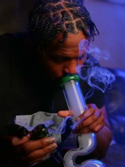 Free A man with dreadlocks using a bong for recreational smoking indoors. Stock Photo