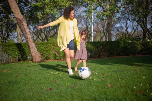 A joyful moment as a mother and daughter play soccer together in a green park setting.