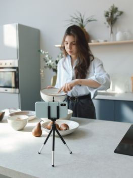 Young woman cooking and filming in a stylish kitchen setup with a smartphone and tripod.