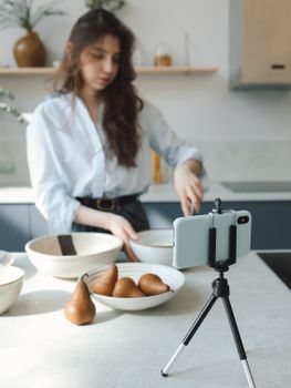 Young woman records cooking tutorial with smartphone on tripod in a bright kitchen.