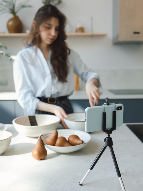 Free Young woman records cooking tutorial with smartphone on tripod in a bright kitchen. Stock Photo