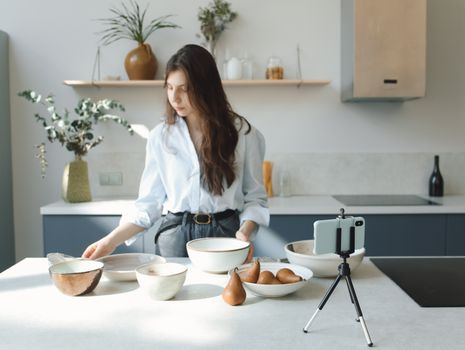 Young woman using smartphone and tripod to record in a modern kitchen with ceramics.