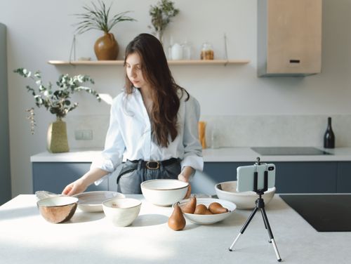 Free Young woman using smartphone and tripod to record in a modern kitchen with ceramics. Stock Photo