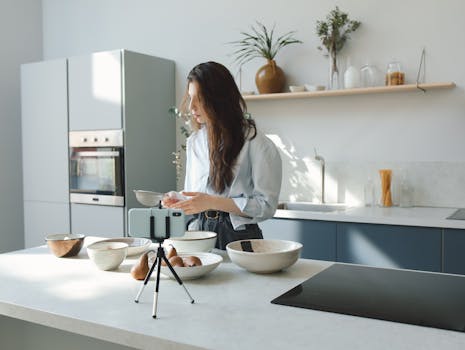 Woman filming a cooking video in a stylish kitchen with a smartphone on a tripod.