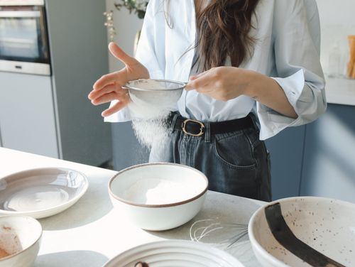 Free A close-up of a woman sifting flour into a bowl in a well-lit modern kitchen. Stock Photo