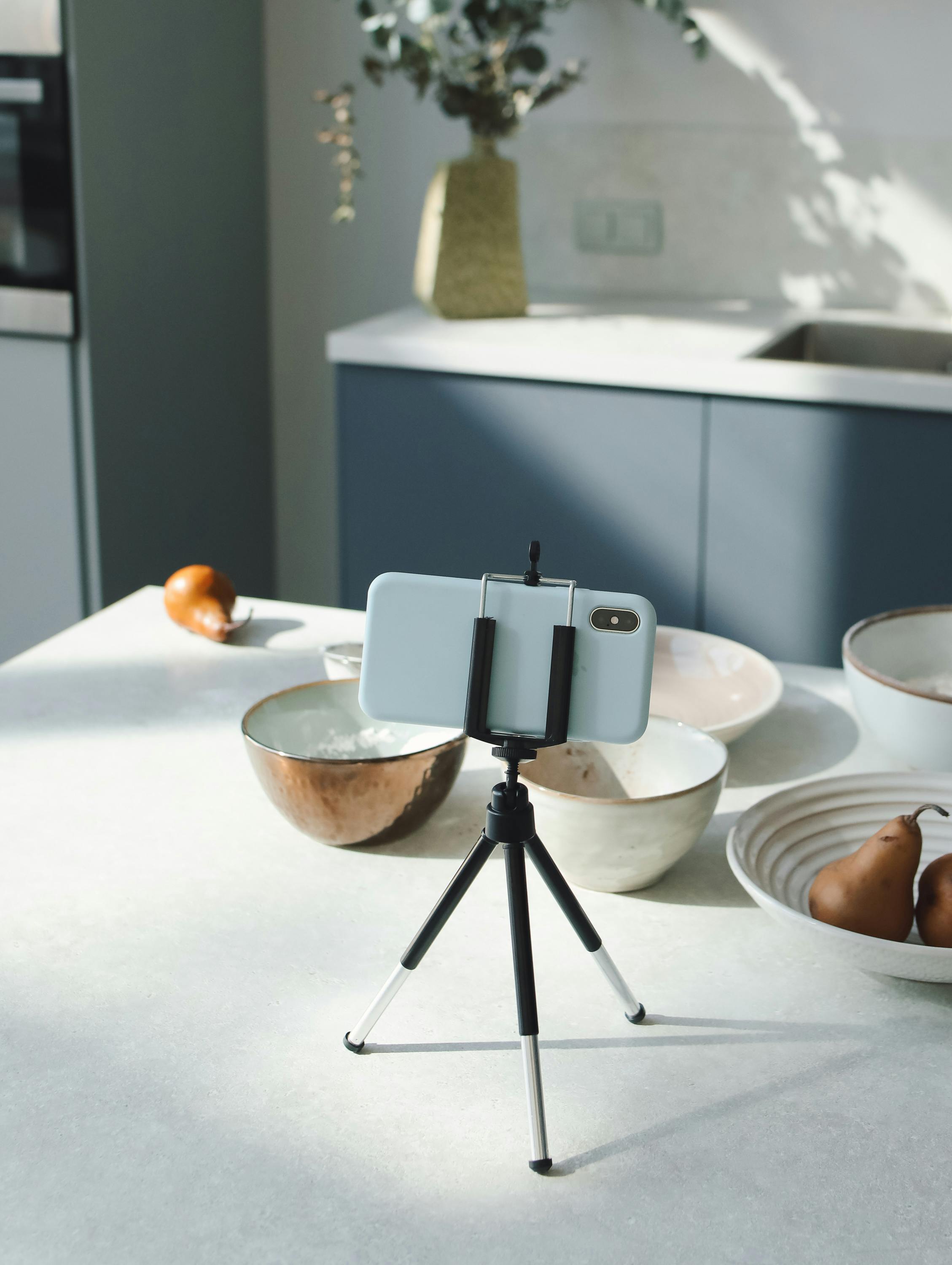 Free A smartphone set up on a tripod in a modern kitchen with bowls and pears, ideal for vlogging or recipe content. Stock Photo