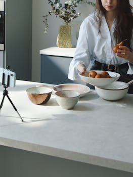 A woman films a food vlog in her kitchen, arranging fruits in bowls for recording.