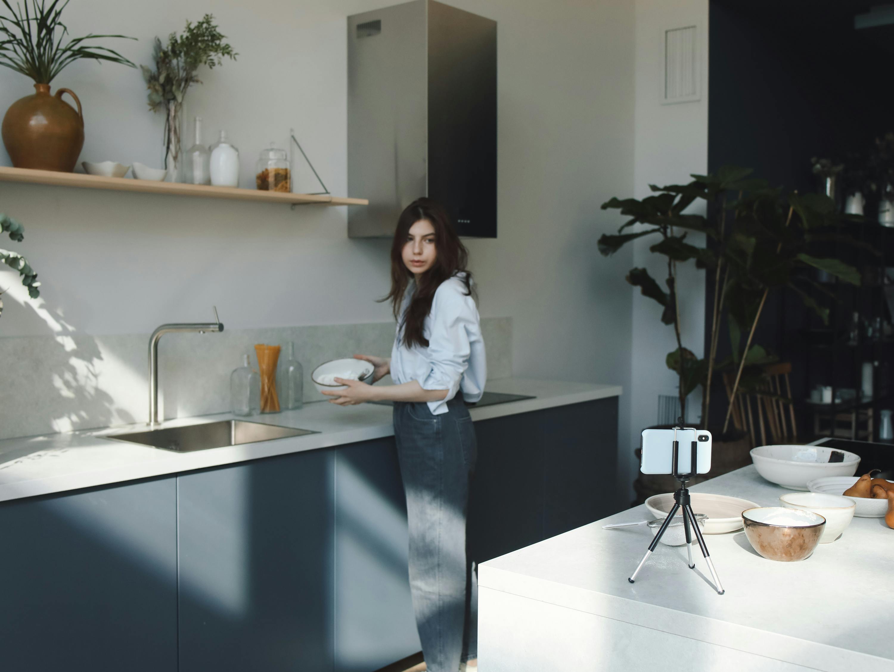 A young woman preparing food in a modern kitchen with smartphone recording.