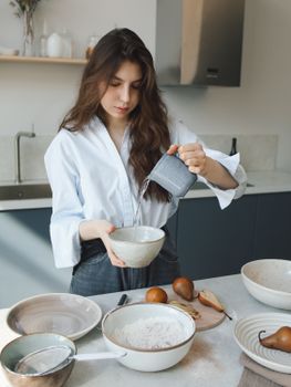 A young woman pours water into a bowl while preparing ingredients for baking in a modern kitchen setting.