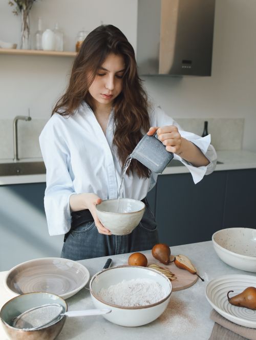 Free A young woman pours water into a bowl while preparing ingredients for baking in a modern kitchen setting. Stock Photo