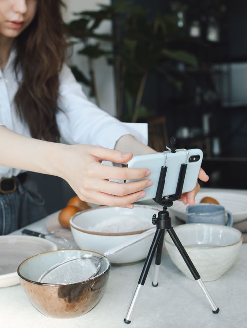 Free A woman arranges a smartphone on a tripod for perfect food photography in a kitchen setting. Stock Photo