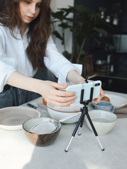 Free Young woman setting up smartphone on tripod to film cooking video indoors. Stock Photo