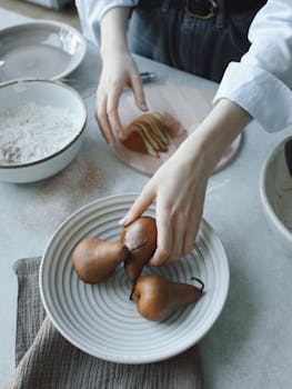 Top view of hands arranging pears for a dessert on a kitchen counter with flour and sliced fruits.