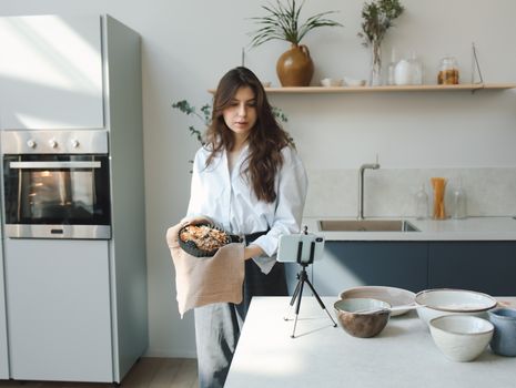 Young woman preparing a dish in a contemporary kitchen, recording with smartphone.