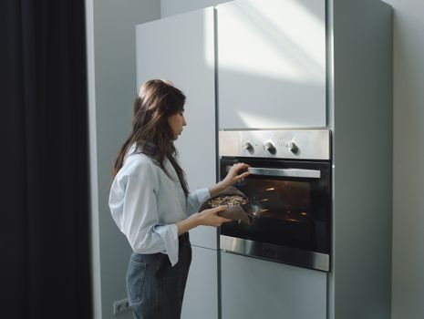 Young woman baking at home, placing a dish in the oven. Bright natural lighting in a modern kitchen setting.