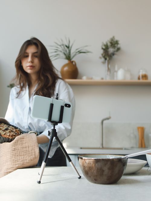 Free Woman vlogging her cooking process in a stylish kitchen using a smartphone on a tripod. Stock Photo