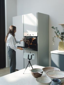 Woman opening oven in a modern kitchen while filming with tripod.
