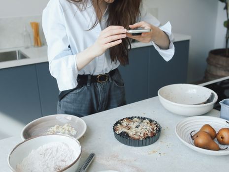 Woman taking a smartphone photo of a homemade pear tart in a modern kitchen setting.